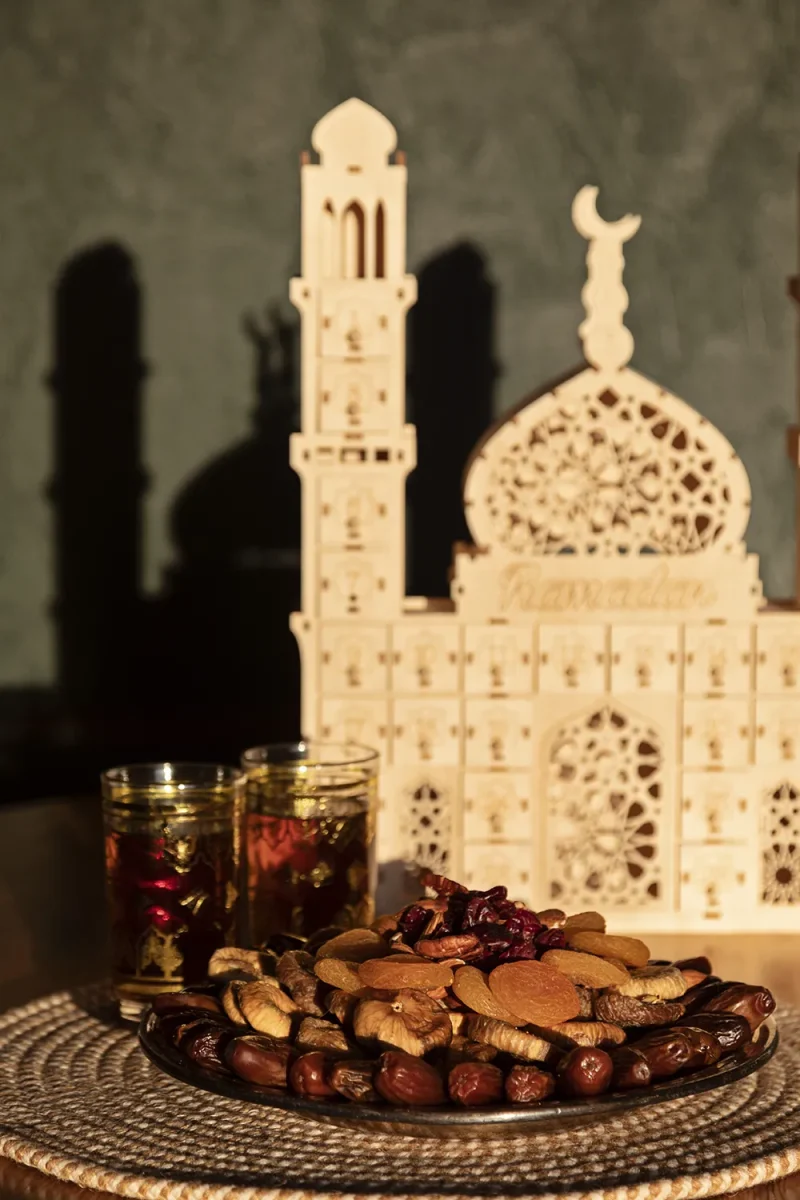 Ahşap cami maketi silueti önünde hurma ve kuru meyveli tabak, “umre hatırası hurma”nın Ramazan ve iftar sofralarına kattığı bereketi vurgularken, hediye kutularına ilham veriyor.

